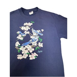 May include: Navy blue t-shirt featuring a detailed illustration of blue jays perched on flowering branches with white and pink blossoms. The word "Jayco" is printed in green below the birds.