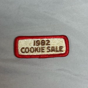 Könnte beinhalten: Gestickter Aufnäher mit rotem Rand und cremefarbenem Mittelteil. Der Text "1982 COOKIE SALE" ist mit braunem Faden gestickt. Der Aufnäher ist rechteckig mit abgerundeten Ecken.