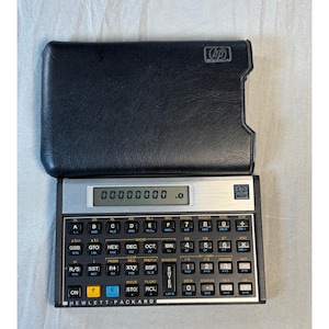 Puede incluir: Una calculadora Hewlett-Packard 16C negra con placa frontal plateada, pantalla digital que muestra ceros y una funda protectora negra de aspecto cuero con el logo HP. El dispositivo tiene numerosas teclas negras con etiquetas blancas.