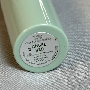 May include: A light mint green lipstick tube with a white circular label on the base. The label reads DIFFERENT LIPSTICK, ROUGE À LÈVRES DIFFÉRENT, ANGEL RED, Special Size/Format Spécial, MADE IN U.S.A., POIDS NET WT. .14 OZ./4 g, CLINIQUE LABS.