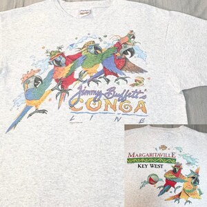 Puede incluir: Camiseta gris claro con un gráfico colorido de loros con sombreros y el texto "Jimmy Buffett's CONGA LINE". Una imagen más pequeña en la parte inferior derecha muestra loros y el texto "Margaritaville Key West".