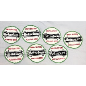 Peut inclure: Ensemble de sept dessous de verre ronds Heineken. Chaque dessous de verre présente le logo Heineken en noir et blanc, avec les mots "IMPORTED" et "HOLLAND BEER" en texte noir. Les dessous de verre ont une bordure verte.