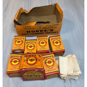 May include: A collection of Hoppe's gun cleaning patches in orange and red cardboard boxes, including a large open display carton. The boxes feature black text, a yellow target graphic, and white fabric patches are stacked to the right side.