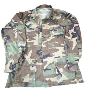 May include: A long-sleeve button-up shirt in a woodland camouflage pattern featuring shades of green, brown, and black. The shirt includes four front pockets with button flaps, a collar, and black text patches reading DUVAL and U.S. ARMY.