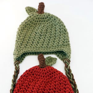 Può includere: Un cappello all'uncinetto fatto a mano a forma di mela. Il cappello presenta una mela rossa con un gambo e una foglia verdi e un cappello verde con lacci intrecciati marroni. Il cappello è progettato per un bambino o un bambino piccolo.