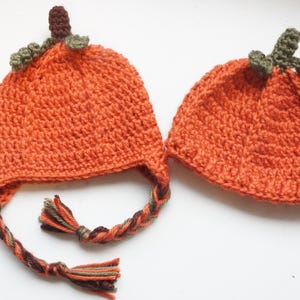 May include: Two orange crochet pumpkin hats. One has ear flaps with braided ties and tassels. The other is a beanie. Both have green leaves and brown stems. Ideal for autumn or Halloween celebrations.