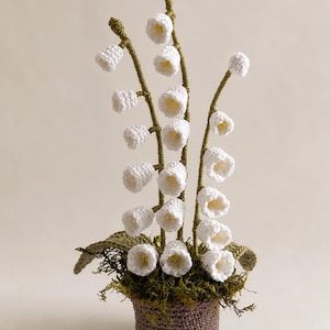 Peut inclure: Une plante muguet au crochet composée de multiples fleurs blanches en forme de cloche sur de fines tiges vertes. La plante présente des feuilles vertes texturées à la base, nichée dans un pot en crochet marron rempli de mousse verte foncée.