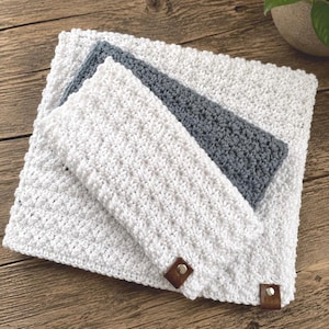 May include: A stack of three crocheted items: a large white square, a smaller gray rectangle, and a white rectangle with brown leather tags. The items are made of a textured, looped yarn, suggesting a handmade craft. The background is a wooden surface.