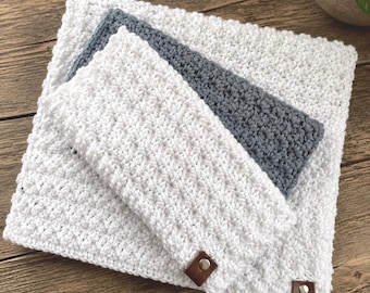 May include: Three crocheted items in a stack: a large white square, a smaller grey rectangle, and a white rectangle with brown leather tags. The items are made of a textured, looped yarn, suggesting a handmade craft. The background is a wooden surface.