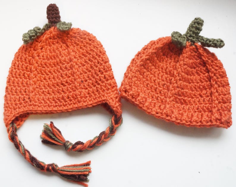Two orange crochet pumpkin hat pattern samples with green leaf stems and braided ear-tie tassels, laid flat on a white background