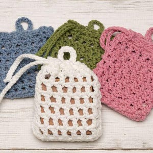 May include: A collection of handmade crochet soap savers in various colors. The set includes a blue, green, pink, and white bag. The white bag has a drawstring closure and a loop for hanging. The bags are made of a textured, open weave.
