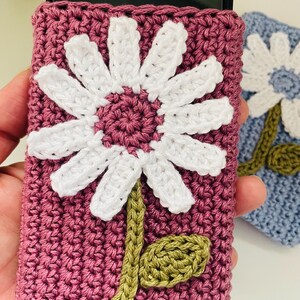 May include: A dark pink crochet phone sleeve featuring a large white daisy with a dark pink center and a green stem with a single leaf. The sleeve is rectangular and textured, designed to hold a smartphone securely within its crocheted fabric.