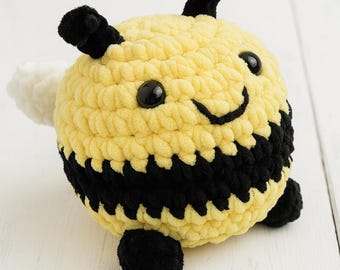 Puede incluir: Un juguete de abeja de peluche hecho a mano. La abeja es principalmente amarilla con rayas negras, antenas negras y pies negros. Tiene una cara sonriente con ojos negros y un ala blanca. El juguete está hecho de hilo suave.