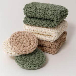 May include: A collection of folded washcloths in green, cream, and brown, with two matching round coasters. The items are made from a thick, woven material. The image is set against a plain background.