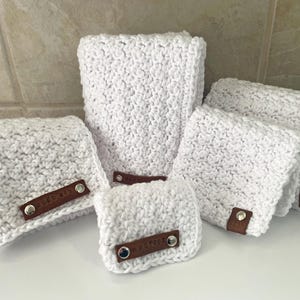 May include: A collection of five white crocheted fabric pouches of varying sizes, each featuring a brown leather-like tab secured with two silver-toned metal rivets. The crochet pattern is textured and consistent across all items in the set.