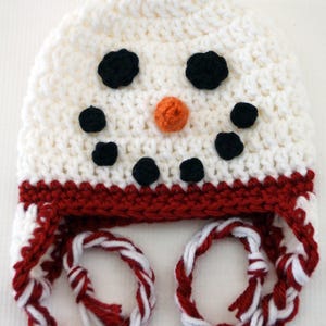 May include: A handmade crochet hat in the shape of a snowman. The hat is white with black dot eyes and mouth, and an orange nose. The brim is red, and the ear flaps have red and white twisted cords.