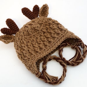 May include: A handmade brown crochet hat shaped like a moose. The hat features dark brown antlers and ears, with braided ties in a lighter brown. The hat is designed for a baby or toddler.