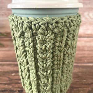 Peut inclure: Un manchon de tasse à café en crochet vert clair avec un motif côtelé. Le manchon est enroulé autour d'une tasse à café réutilisable bleu clair avec un couvercle blanc. L'arrière-plan est une surface en bois.