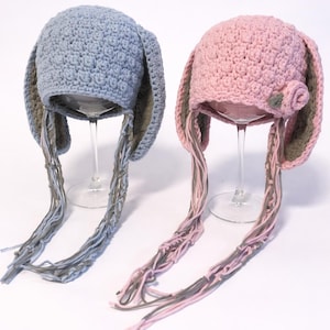 May include: Two handmade crochet bunny hats. One is light blue with gray trim and dangling strands. The other is pink with green trim and a small flower accent. Both are displayed on clear glass stands.