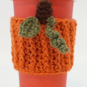 Puede incluir: Una taza de café de papel roja con tapa blanca y un portavasos de calabaza de ganchillo naranja hecho a mano. El portavasos presenta un tallo marrón y hojas verdes, perfecto para el otoño.