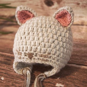 May include: A beige crocheted baby hat shaped like a cat, featuring pink inner ears and brown facial details. The hat has ear flaps with braided ties.