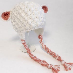 May include: A white crochet hat with pink accents, featuring ear-shaped details and braided ties. The hat is handmade and suitable for children. The braids are a mix of white and pink yarn.