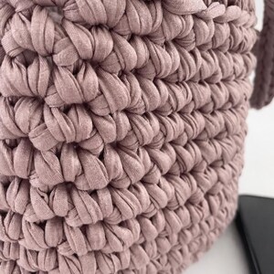 May include: Close-up of a dusty rose-colored crochet bag. The bag is made of thick, textured yarn, with a tightly woven pattern. The handle is visible on the right side. The bag is sitting on a white surface.