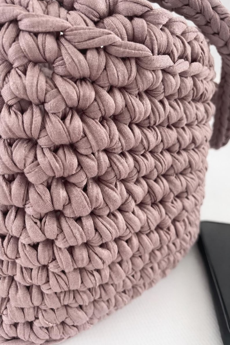 May include: Close-up of a dusty rose-colored crochet bag. The bag is made of thick, textured yarn, with a tightly woven pattern. The handle is visible on the right side. The bag is sitting on a white surface.