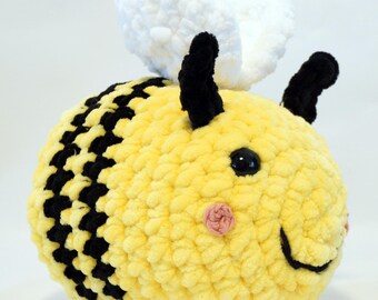 Puede incluir: Un juguete de abeja de peluche hecho a mano, con cuerpo amarillo y rayas negras. La abeja tiene antenas negras, una cara sonriente y alas blancas. El juguete está hecho de hilo suave y texturizado.