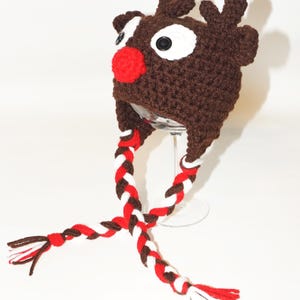 Op de afbeelding: Een bruine gehaakte hoed in de vorm van een rendier, met witte ogen, een rode neus en kleine geweien. De hoed heeft lange, gevlochten banden in rood, wit en bruin. Een leuk, feestelijk winteraccessoire.