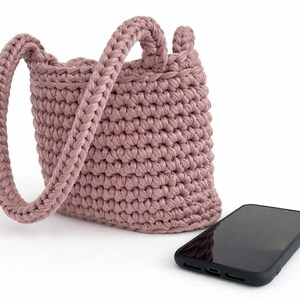 May include: A dusty rose crochet bucket bag with a single braided handle. The bag is next to a black smartphone. The bag's texture is visible, and the handle is thick and sturdy. The bag is likely handmade.