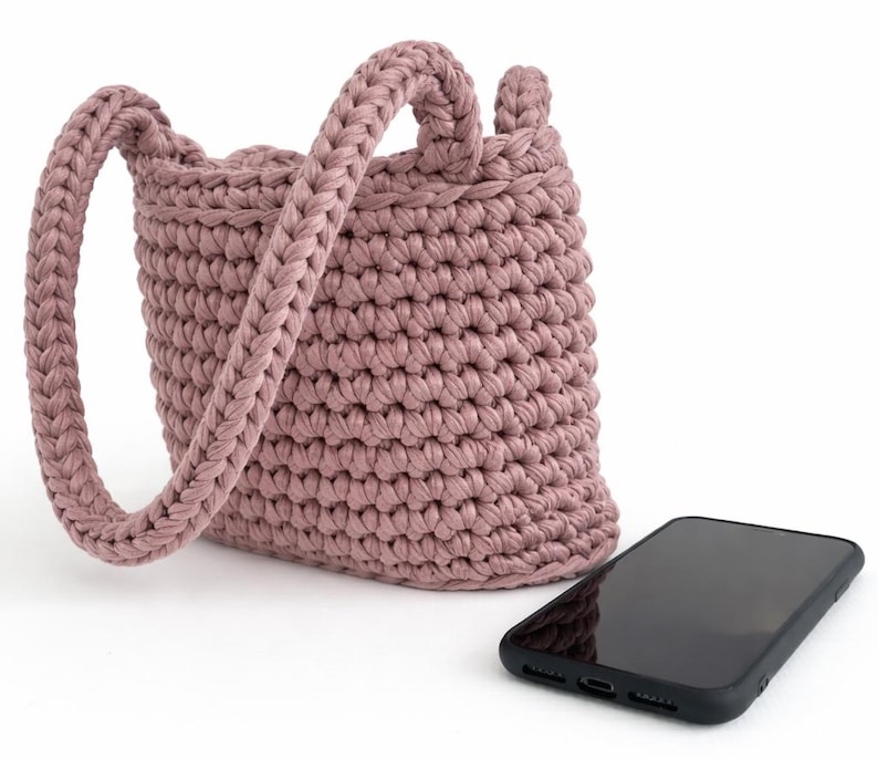 May include: A dusty rose crochet bucket bag with a single braided handle. The bag is next to a black smartphone. The bag's texture is visible, and the handle is thick and sturdy. The bag is likely handmade.