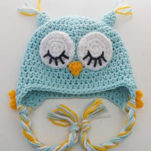 May include: A light blue crochet owl hat with white eyes, black eyelashes, and a yellow beak. The hat has ear flaps with yellow, white, and blue braided ties. The top of the hat has small yellow and white tassels.