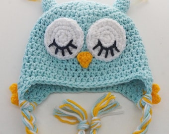 May include: A light blue crochet owl hat with white eyes, black eyelashes, and a yellow beak. The hat has ear flaps with yellow, white, and blue braided ties. The top of the hat has small yellow and white tassels.