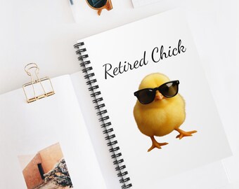 May include: A white spiral-bound notebook featuring the text "Retired Chick" and a cartoon chick wearing black sunglasses. The notebook is open, with a gold-coloured binder clip and sunglasses on a white surface.