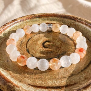 May include: A beaded bracelet featuring round, translucent white and orange-tinted gemstone beads. The bracelet includes a single, cylindrical silver-toned metal spacer bead with an intricate, embossed floral pattern centered at the top.