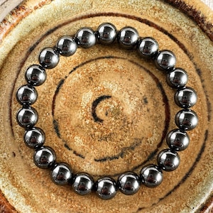 May include: A dark grey beaded bracelet with a reflective, metallic sheen. The bracelet is made of round beads, arranged in a circular pattern. The beads are set against a textured, brown background.
