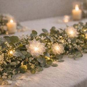 Puede incluir: Un centro de mesa con una guirnalda de vegetación, pequeñas flores blancas y luces de hadas. El arreglo incluye grandes acentos florales iluminados en forma de estrella y velas, creando un ambiente suave y romántico.