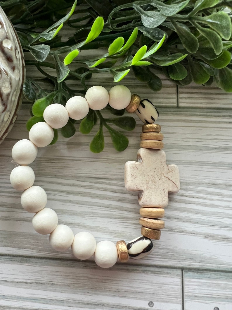 May include: White bead bracelet with a cross charm and gold and black accents.