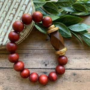May include: A red wooden bead bracelet with a gold and brown wooden accent. The bracelet is on a wooden surface with green leaves.