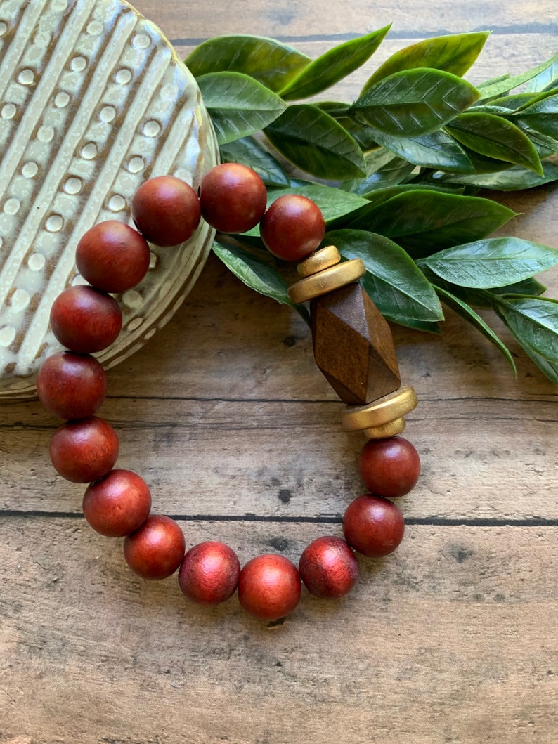 May include: A red wooden bead bracelet with a gold and brown wooden accent. The bracelet is on a wooden surface with green leaves.