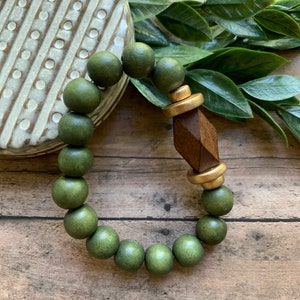 May include: A green wooden bead bracelet with a gold and brown geometric charm. The bracelet is on a wooden surface.