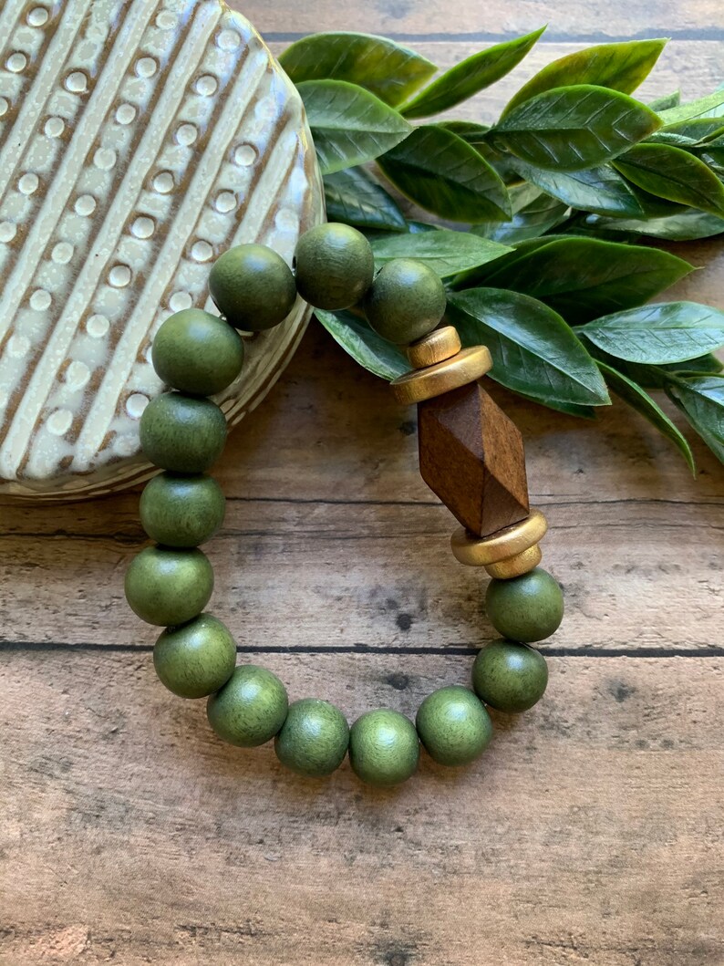 May include: A green wooden bead bracelet with a gold and brown geometric charm. The bracelet is on a wooden surface.