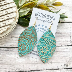 May include: Teal leather earrings with a gold embossed floral pattern. The earrings are shaped like a teardrop and have a gold hook closure. The earrings are displayed on a white card with the text "Beaded Blues Handcrafted Jewelry" and the social media handle "@BeadedBlues".