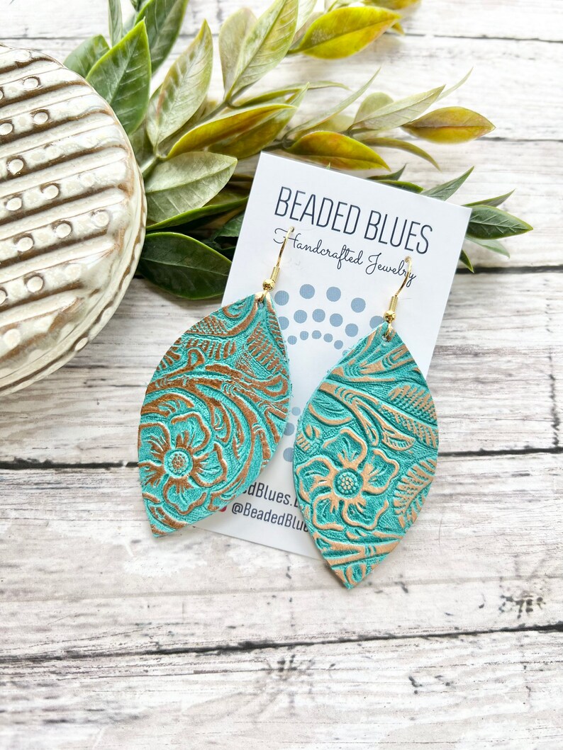 May include: Teal leather earrings with a gold embossed floral pattern. The earrings are shaped like a teardrop and have a gold hook closure. The earrings are displayed on a white card with the text "Beaded Blues Handcrafted Jewelry" and the social media handle "@BeadedBlues".