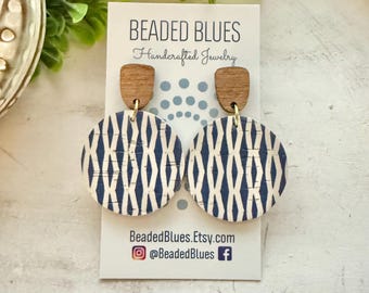 May include: A pair of handcrafted earrings from Beaded Blues. The earrings feature a wooden top and a circular design with a white background and a navy blue geometric pattern. The earrings are displayed on a white card with the brand's name and social media handles.
