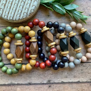 May include: A set of six wooden bead bracelets with geometric wooden charms and gold accents. The bracelets are in shades of green, yellow, red, black, gray, and beige.