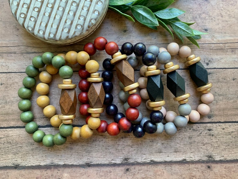 May include: A set of six wooden bead bracelets with geometric wooden charms and gold accents. The bracelets are in shades of green, yellow, red, black, gray, and beige.