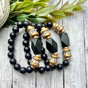 May include: Three black wooden bead bracelets with gold and white patterned beads. The bracelets are stacked on top of each other.