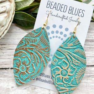 May include: Teal leather earrings with a gold embossed floral pattern. The earrings are shaped like a teardrop and have a gold hook closure. The earrings are handmade and are a beautiful statement piece.